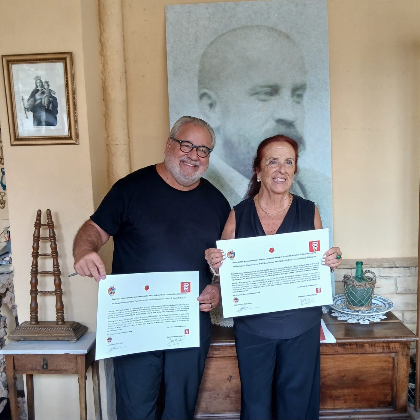 Un hombre y una mujer sonríen mostrando sendos diplomas con sello rojo, de pie ante un gran retrato en blanco y negro de Antoni Gaudí.