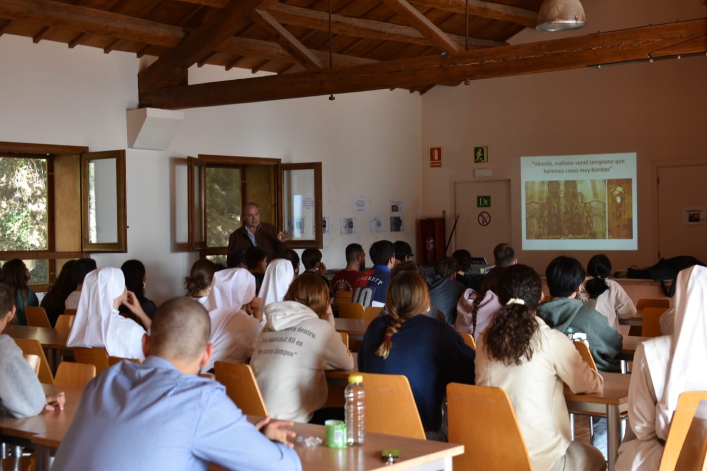 Un hombre de pie junto a una ventana da una charla ante un grupo numeroso de jóvenes sentados en mesas, con una diapositiva proyectada de fondo, en una sala de vigas de madera.