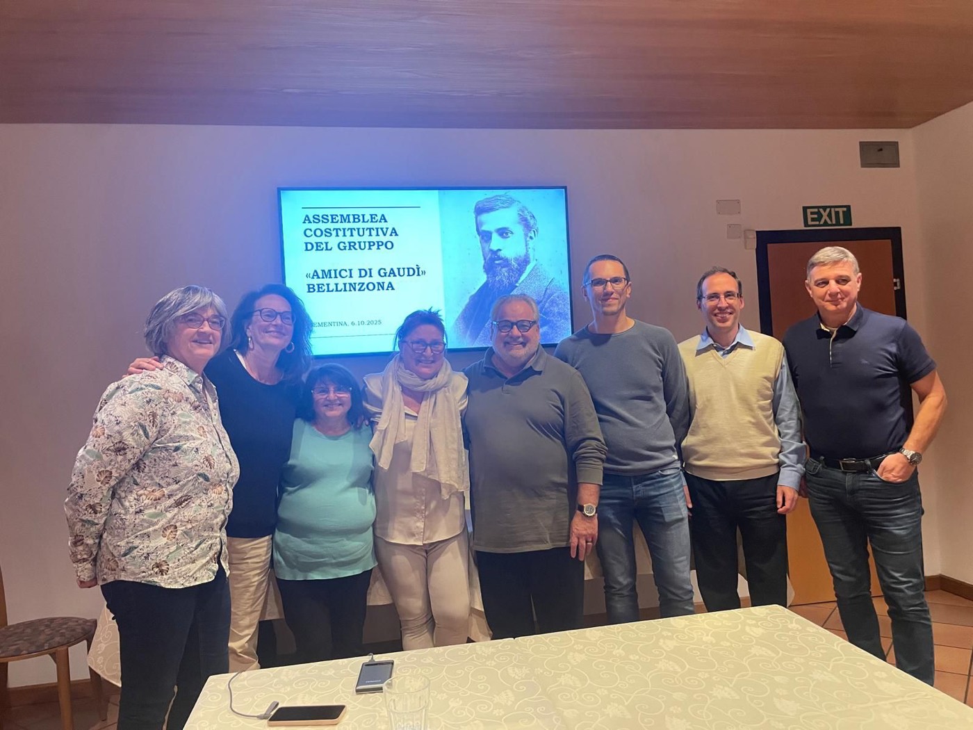 Ocho personas posan sonrientes en una sala junto a una pantalla que muestra el retrato de Gaudí y el título «Assemblea Costitutiva del Gruppo Amici di Gaudì Bellinzona».