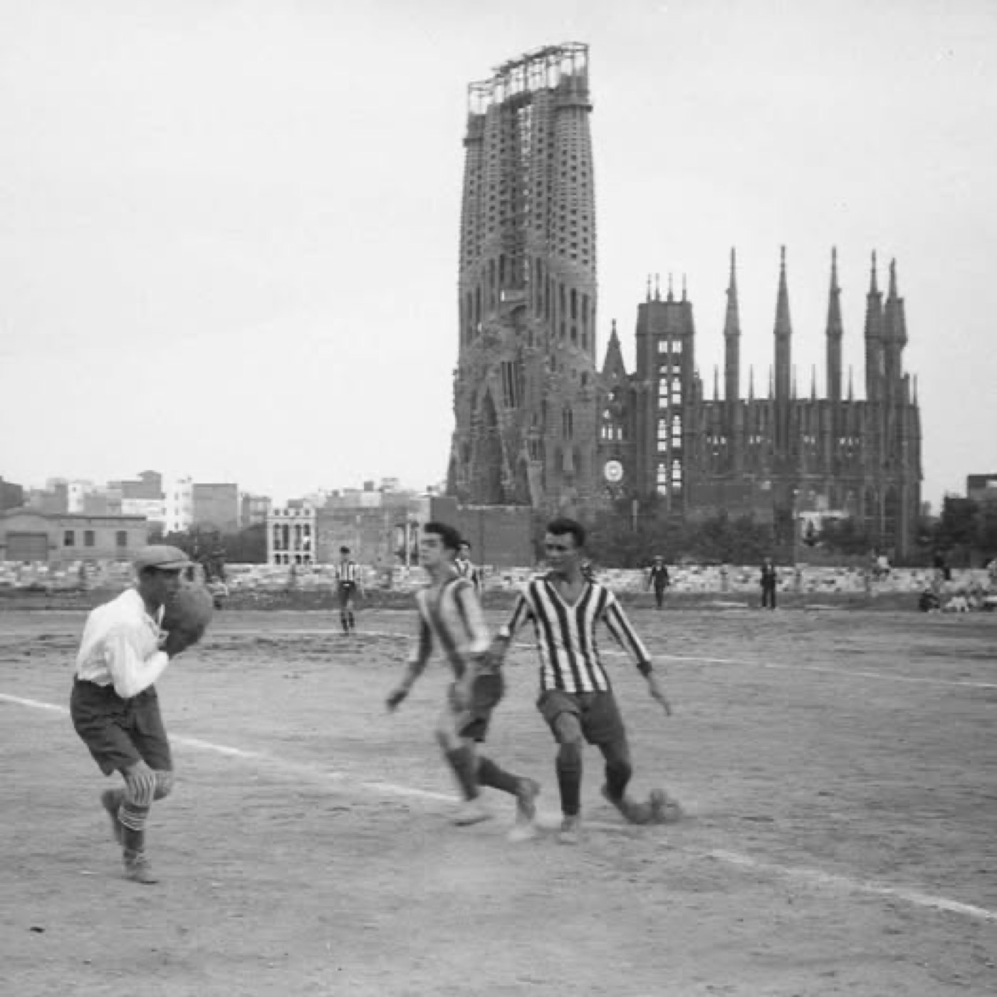Fotografía histórica en blanco y negro de un partido de fútbol callejero con la Sagrada Família, aún en construcción y con solo las torres de la fachada del Nacimiento, al fondo
