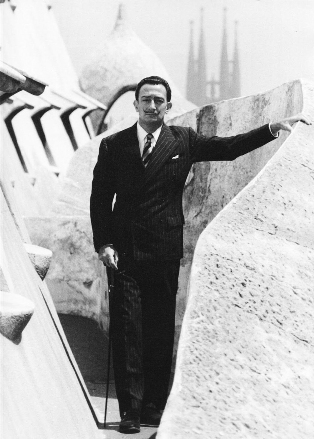 Fotografía histórica en blanco y negro de Salvador Dalí posando con bastón en la azotea de la Casa Milà (La Pedrera), con las torres de la Sagrada Família al fondo