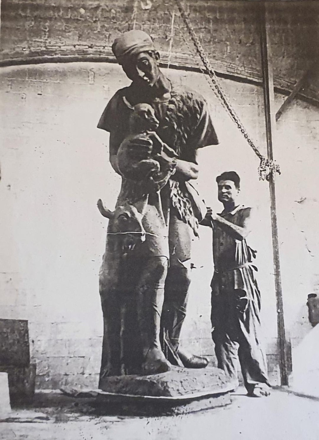 Fotografía histórica en blanco y negro de una gran estatua de un pastor con un cordero en brazos, aún en el taller, junto a un hombre que trabaja en ella.