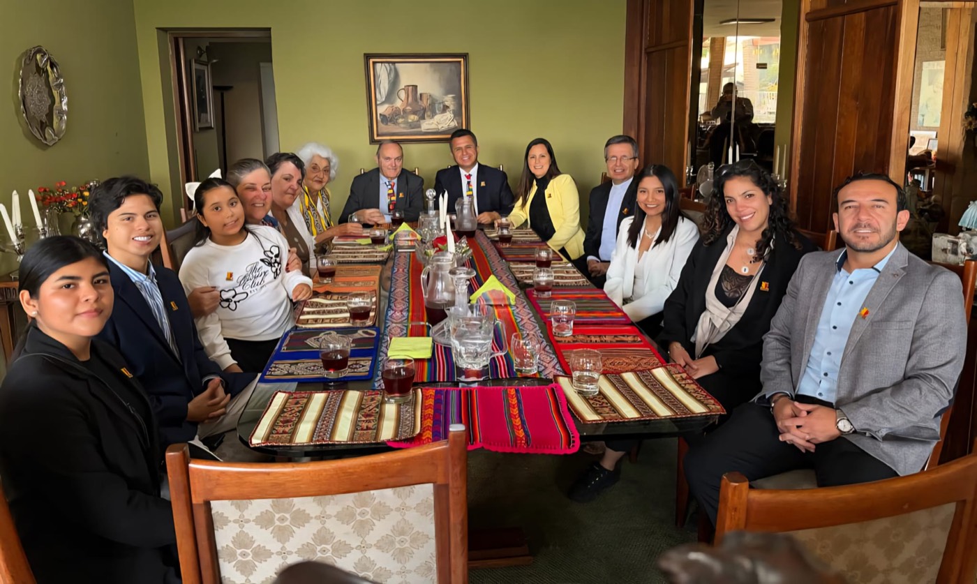 Grupo numeroso de personas reunidas alrededor de una mesa larga con manteles andinos, en una comida de trabajo en un salón.