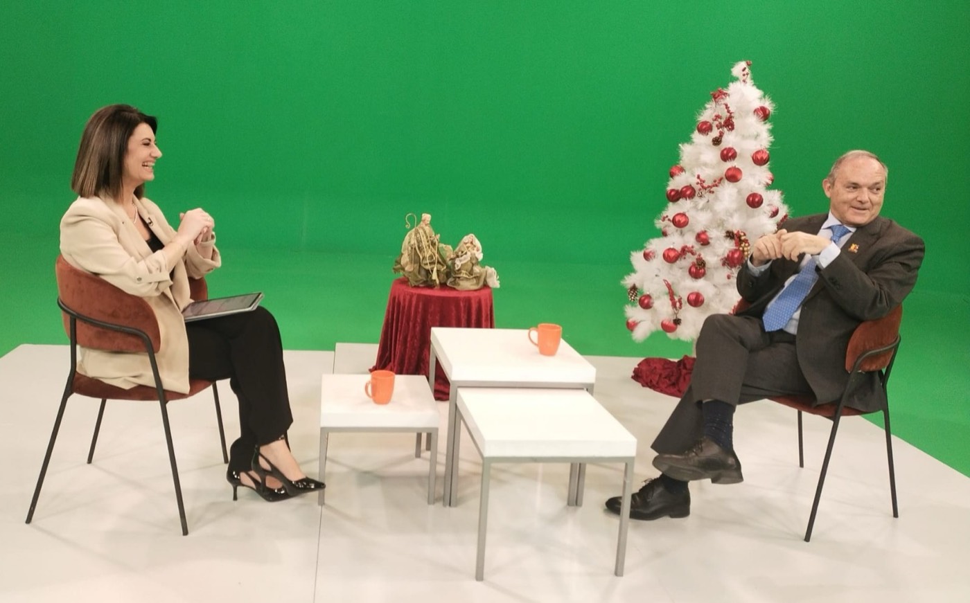 José M. Almuzara, sentado, es entrevistado por una periodista en un plató de televisión con fondo de croma verde, mesas bajas y un árbol de Navidad.