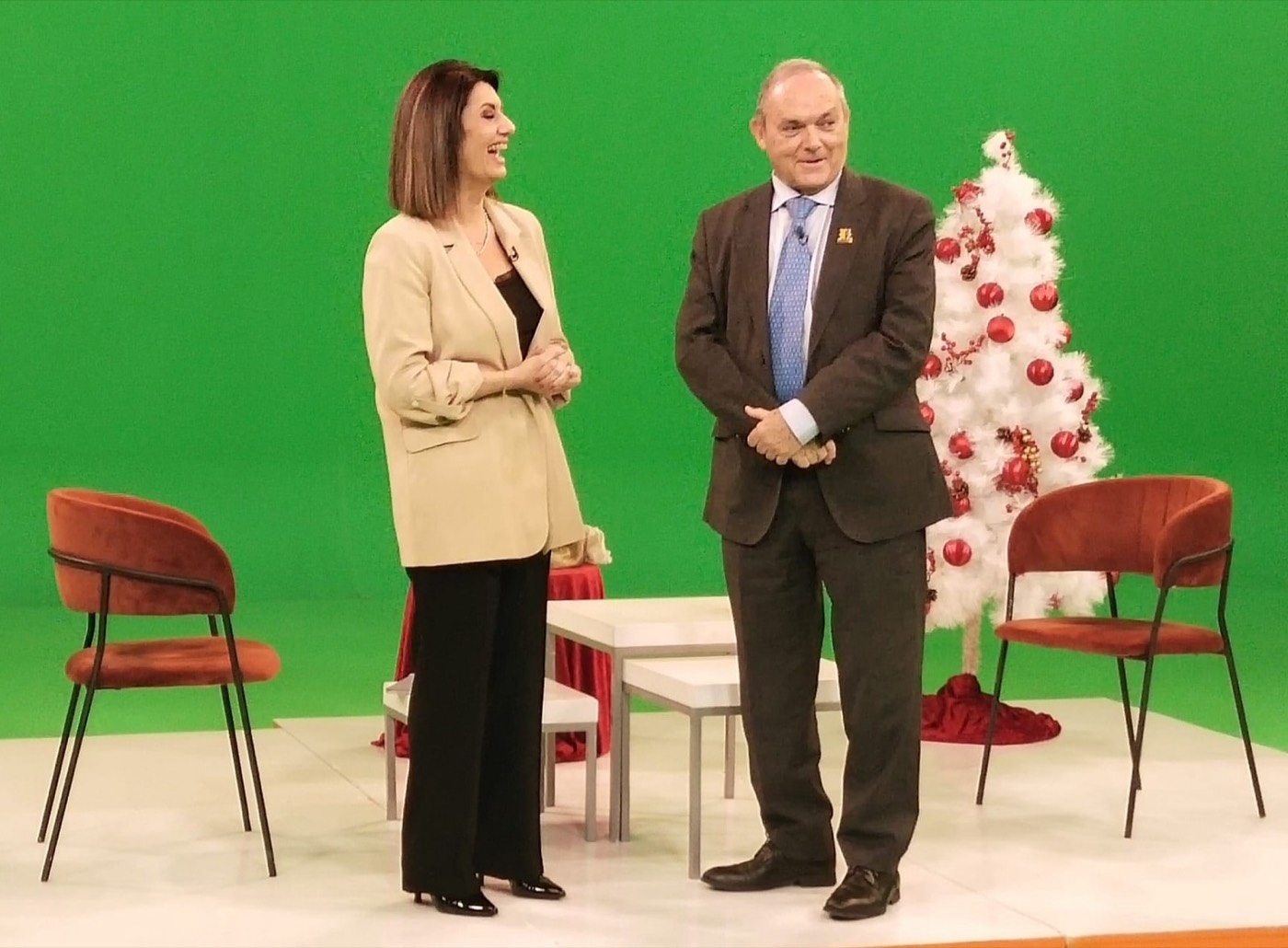 José M. Almuzara conversa de pie con una periodista en un plató de televisión con fondo de croma verde y un árbol de Navidad decorado.