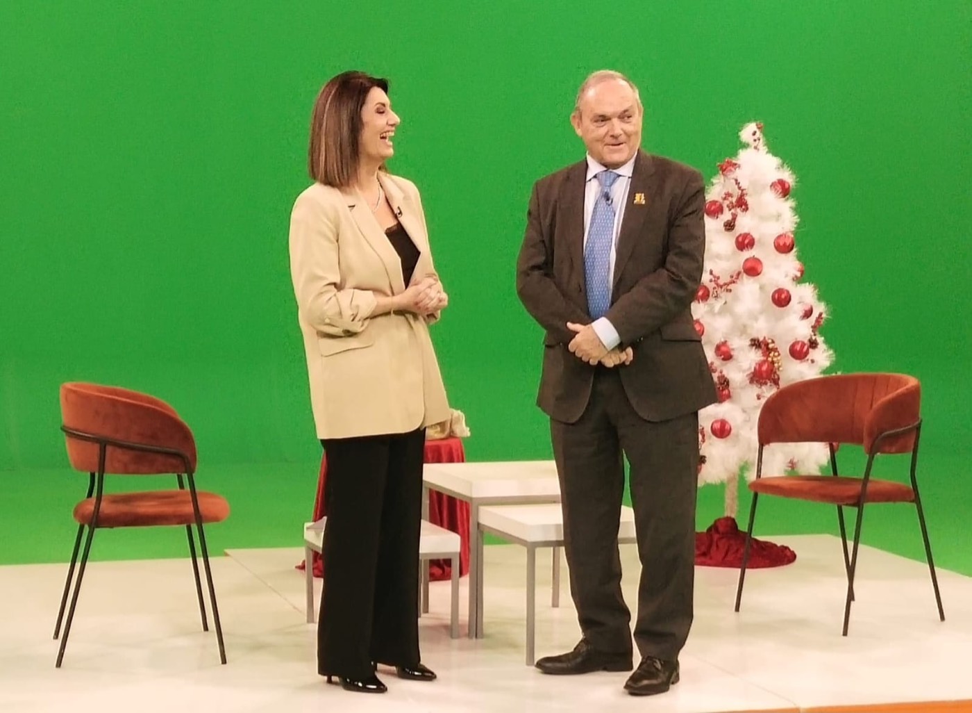 José M. Almuzara conversa de pie con una periodista en un plató de televisión con fondo de croma verde y árbol navideño, en otro momento de la misma grabación.