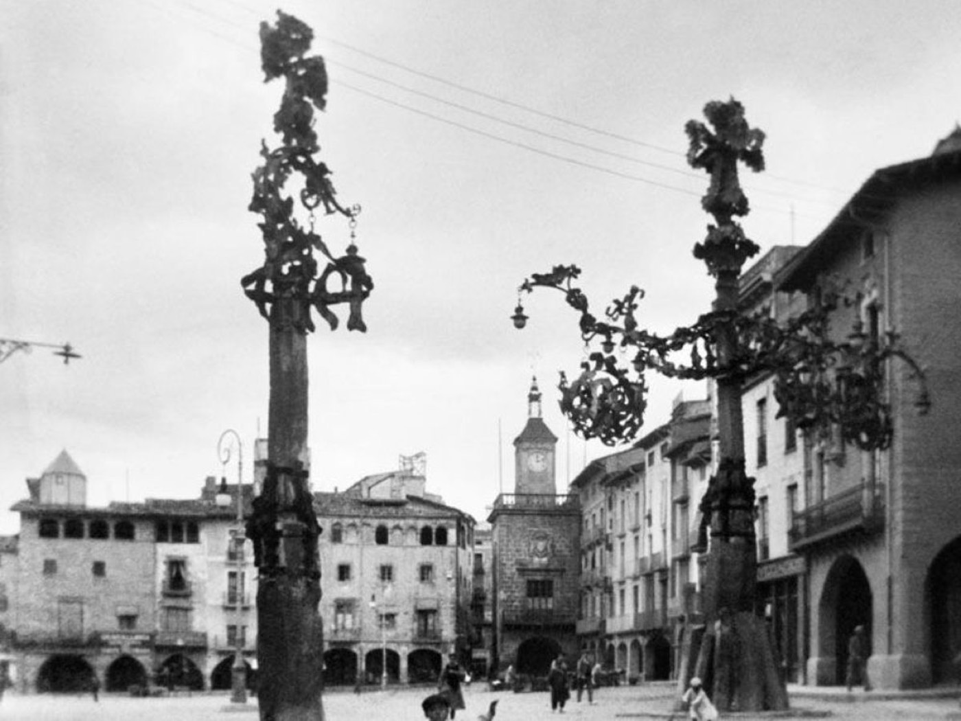 Fotografía histórica en blanco y negro de la plaza Mayor de Vic con las dos farolas modernistas de hierro forjado y piedra diseñadas por Gaudí, hoy desaparecidas, y el campanario del ayuntamiento al fondo.