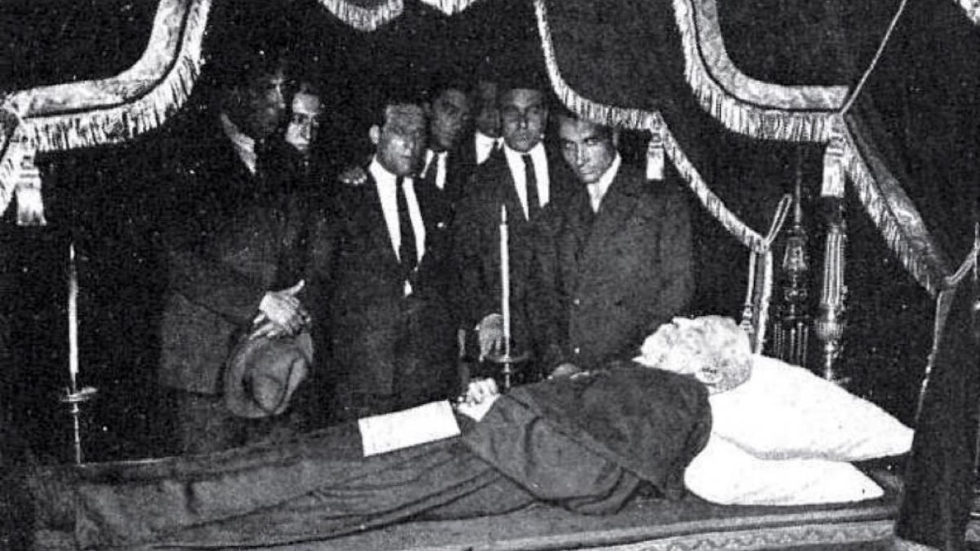 Fotografía histórica en blanco y negro de la capilla ardiente de Antoni Gaudí, con varios hombres de luto junto a su cuerpo yacente.