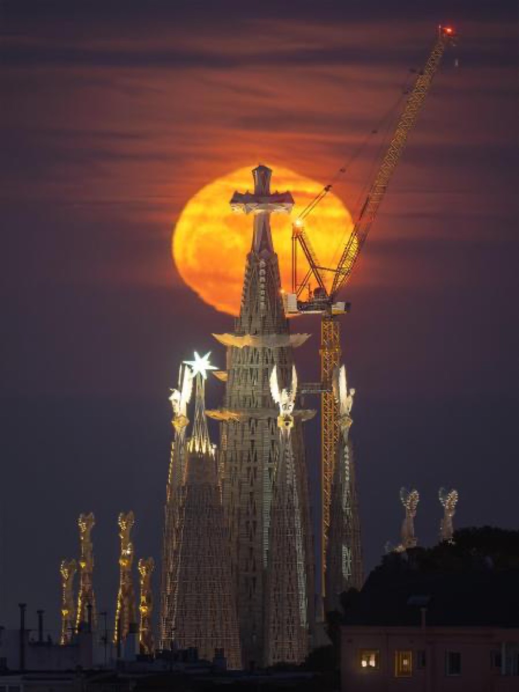 La luna llena de color naranja se eleva justo detrás de la cruz de las torres centrales de la Sagrada Família, iluminada de noche, con una grúa de obra al fondo.