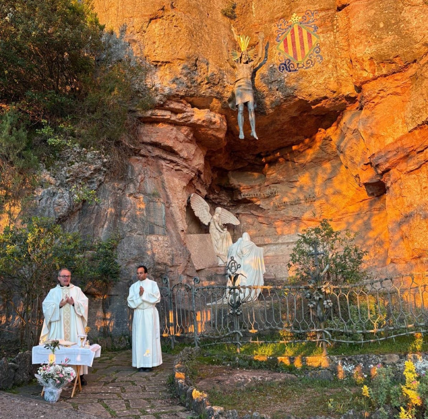 Altar preparado con flores ante la hornacina rocosa del Primer Misterio de Gloria de Montserrat (la Resurrección, obra de Gaudí), con dos sacerdotes vestidos de blanco de pie junto a él al atardecer.