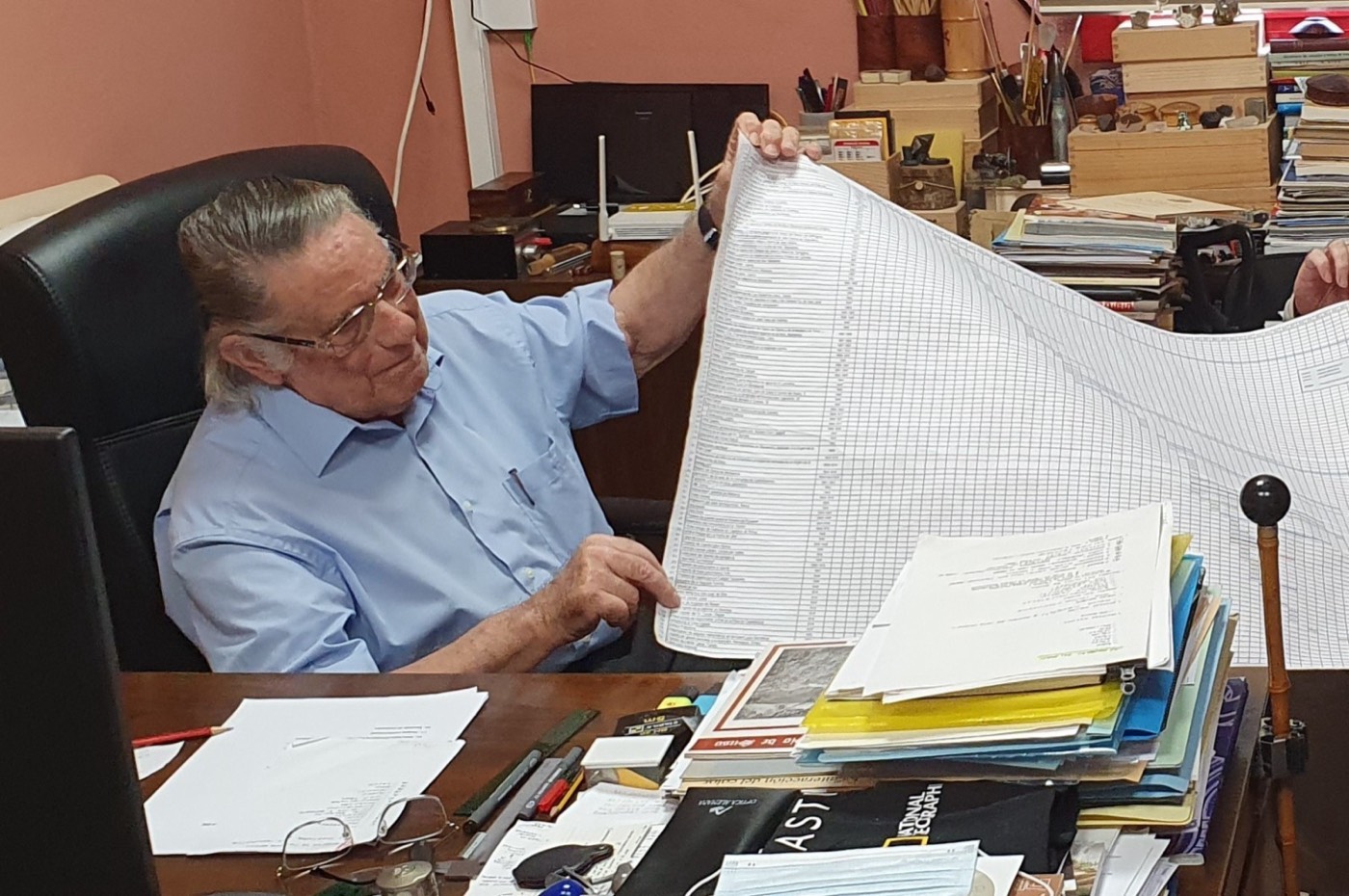 Un hombre mayor de pelo largo y canoso, sentado en su despacho, sostiene y revisa un extenso listado impreso rodeado de libros y documentos.