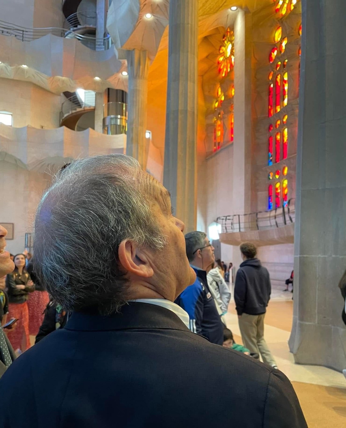 Un hombre visto de perfil, con el pelo cano, contemplando los vitrales de colores y las columnas del interior de la Sagrada Família, rodeado de otros visitantes.