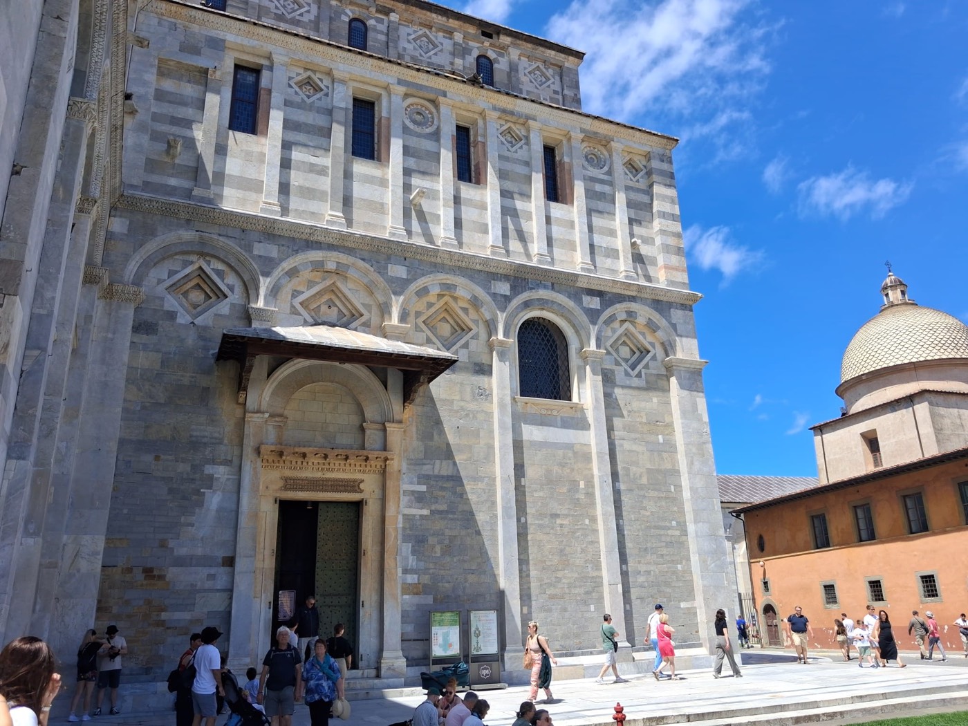 Fachada lateral de piedra de la Catedral de Pisa con una puerta de acceso, con gente entrando y saliendo.