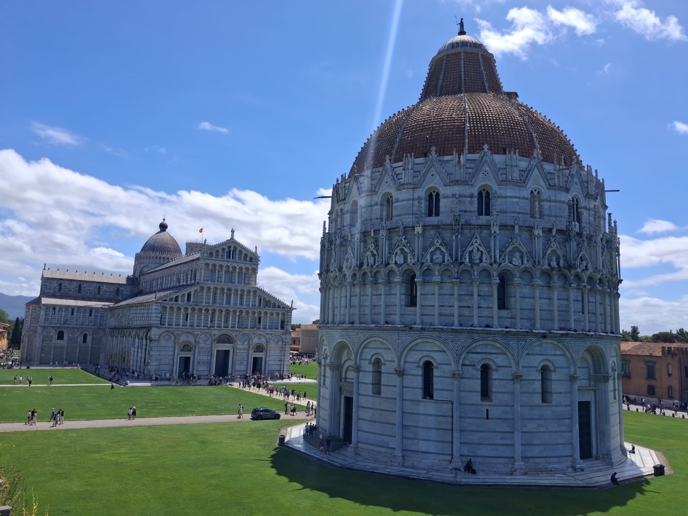 Vista cercana del Baptisterio de Pisa con su cúpula, y la Catedral al fondo, bajo un cielo azul con nubes.