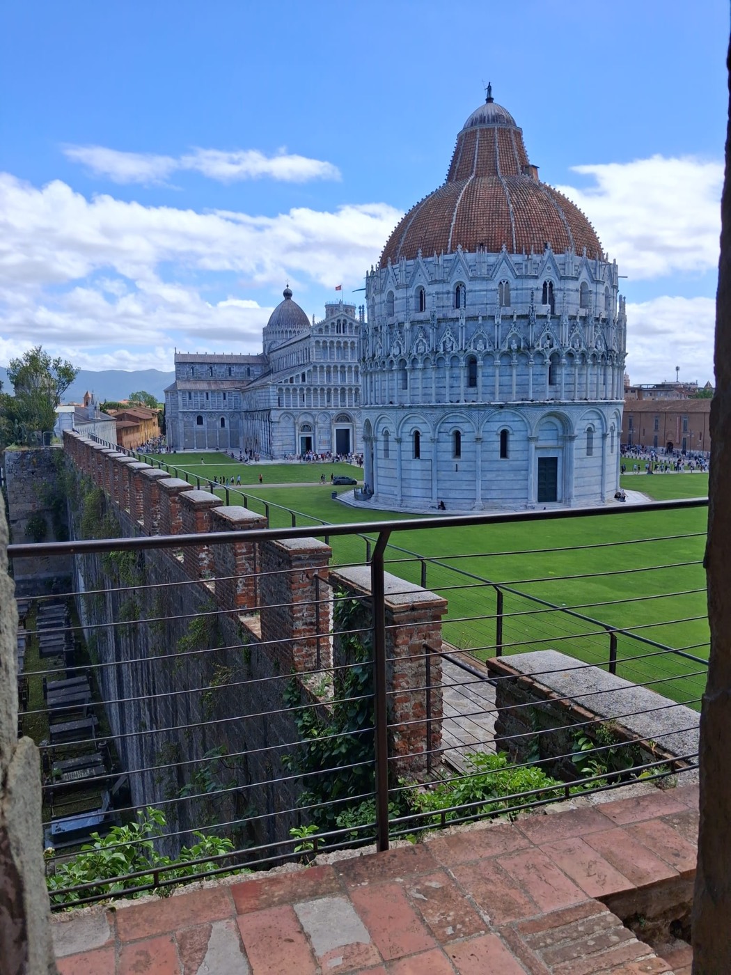 Vista del Baptisterio y la Catedral de Pisa desde lo alto de la muralla, con una barandilla y columnas de ladrillo en primer plano.