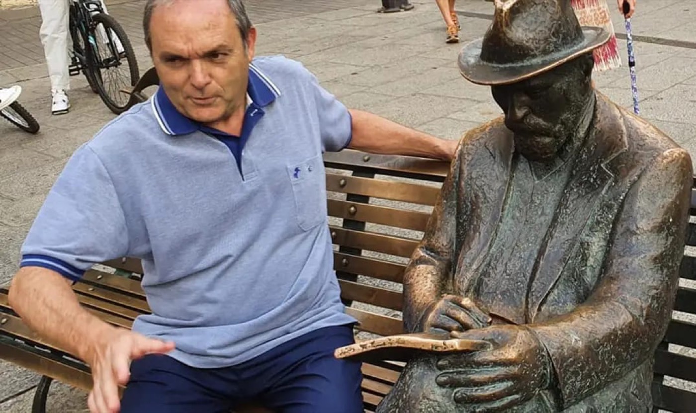 Un hombre sentado en un banco de una calle peatonal junto a una escultura de bronce de Antoni Gaudí sentado con sombrero.