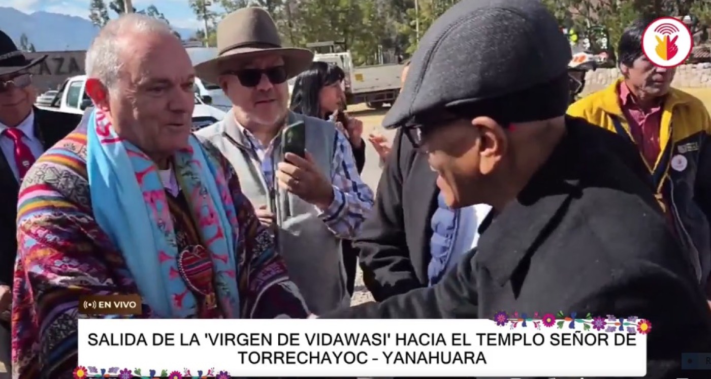 José Manuel Almuzara, con poncho tradicional andino, saluda de la mano a un hombre mayor durante la procesión de la Virgen de Vidawasi, en una captura de una emisión en directo.