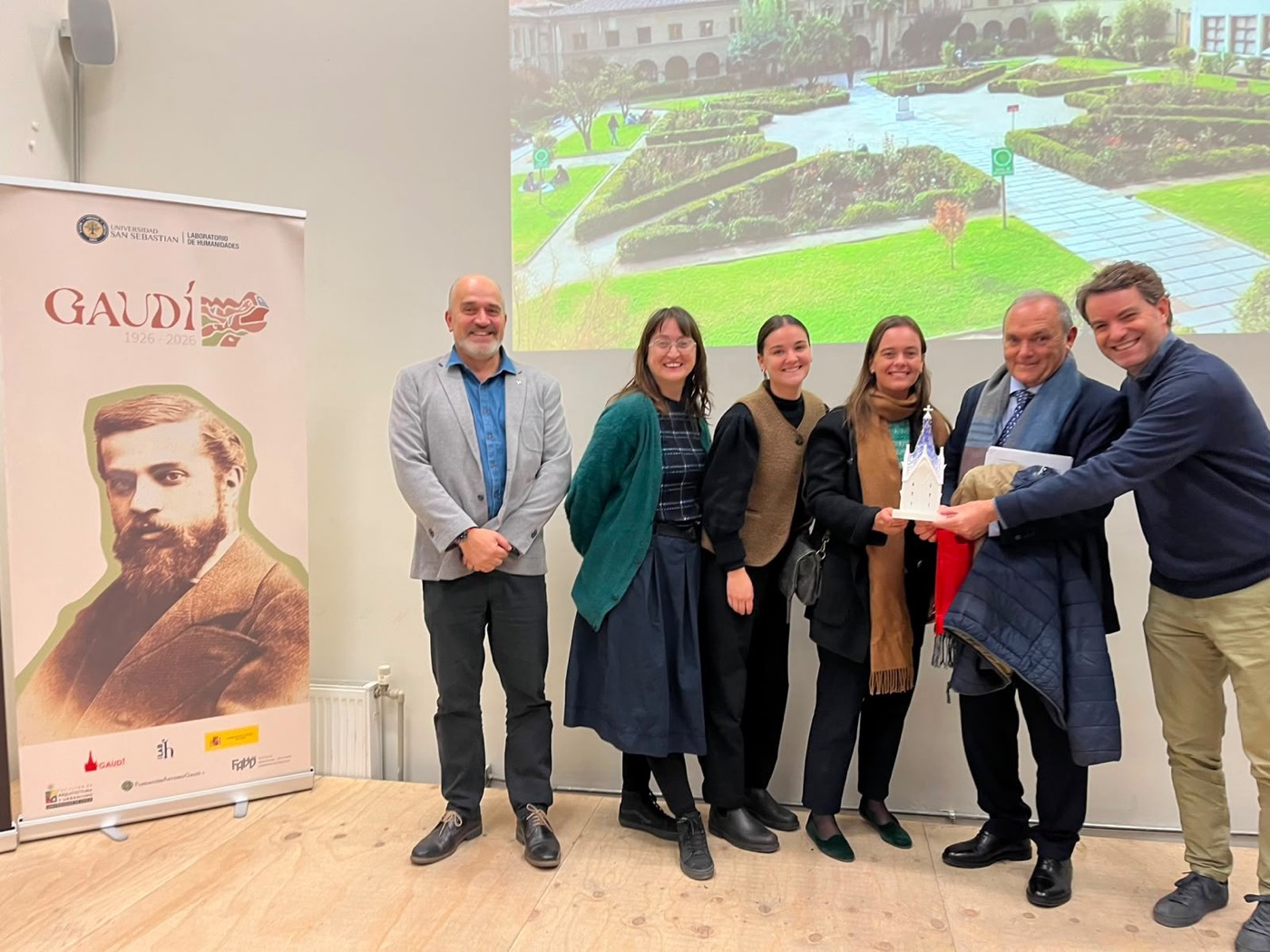 José Manuel Almuzara y un grupo de profesores y estudiantes posan sonrientes junto a un roll-up de 'Gaudí' en la Universidad San Sebastián de Santiago de Chile, sosteniendo una pequeña figura conmemorativa.