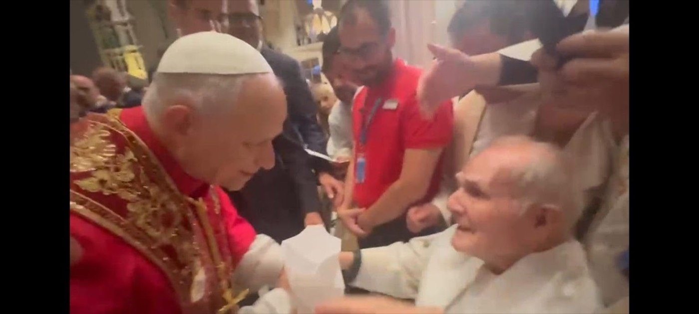 El papa, vestido de rojo, bendice a un sacerdote anciano sentado en silla de ruedas, rodeado de varias personas.